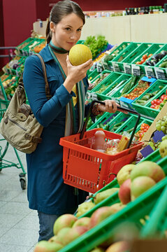 Woman smelling a melon in a grocery store produce section, holding a basket in her hand. Germany