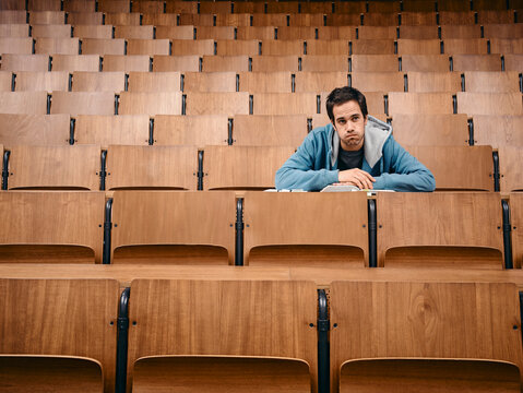 A young man sits alone in a wooden auditorium with empty seats surrounding him. Germany
