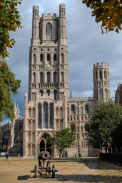 Historic cathedral with stone towers, surrounded by trees and a cannon in the foreground. Ely, England