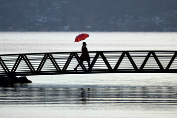 Silhouetted person with a red umbrella crosses a bridge over calm water at dusk. Bainbridge Island, WA, USA