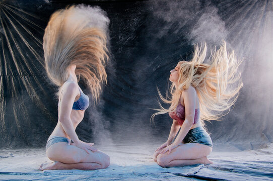 Two women kneel on a textured surface with hair flipping dramatically against dark backdrop. WA, USA