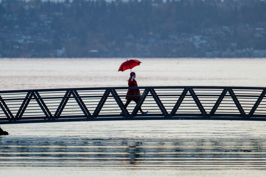 A person with a red umbrella walks across a metal bridge over calm water. Bainbridge Island, WA, USA