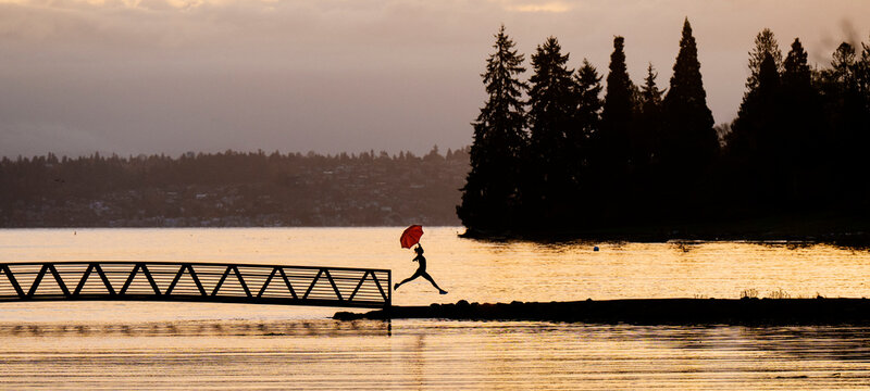 Silhouette of a person with a red umbrella jumping on a bridge near a lake at sunset. Bainbridge Island, WA, USA