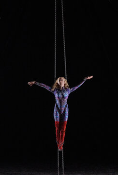 Aerial acrobat performs suspended in chains wearing a colorful bodysuit and red boots. WA, USA
