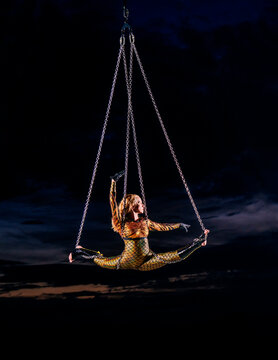 Performer in a gold costume suspended in mid-air by chains against a dark sky. WA, USA
