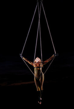 Performer in a gold costume suspended in mid-air by chains against a dark sky. WA, USA