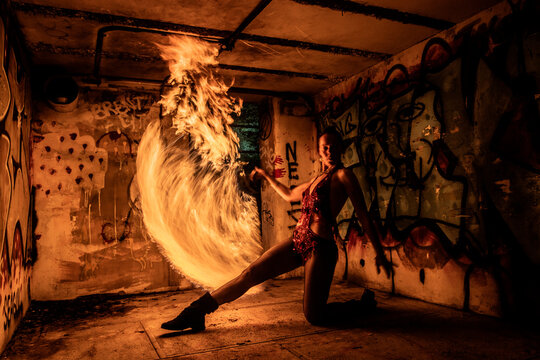 Fire performer kneeling, spinning fire poi in a graffiti-covered room at night.  WA, USA