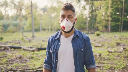 Caucasian male volunteer wearing respirator mask. Bearded man standing alert and focused. Denim shirt over white T-shirt. Forest clearing around him. Preparing environmental cleanup work outdoors.