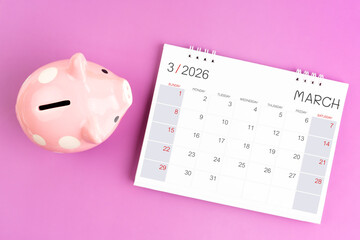 Desk calendar for March 2026 and pink piggy bank on purple background.