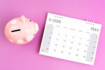 Desk calendar for May 2026 and pink piggy bank on purple background.