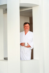 Adult Blond Man in White Robe Standing by the Window Enjoying a Warm Cup of Coffee, Embracing a Peaceful Morning at Home.