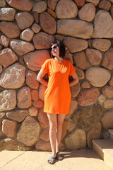 Elegant woman orange dress standing against rustic stone wall, wearing sunglasses outdoors.