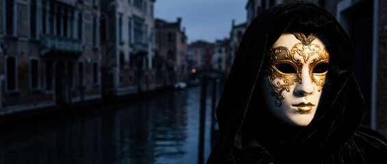 Mysterious figure wearing an elegant gold and white Venetian mask and black hooded cloak by a dark canal in Venice at dusk. Carnival of Venice masquerade concept.