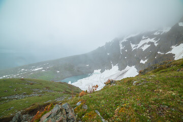 Scenic misty foggy top view from green grassy slope to frozen lake among rocks and snows in mountain cirque in rain. Ice floes float on turquoise water of alpine lake under cloudy sky in rainy weather