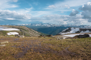 Field of thawed ground with old grass and marsh among rocks and snowfields on hill top with view to big snowy mountain range far away under cloudy sky. Sunlight and shadow of clouds on forest valley.