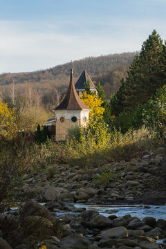 Country house is located on banks of mountain river. Octagonal tower with pointed roof and round windows, partially hidden by lush foliage, rises above autumn trees and is surrounded by cliffs