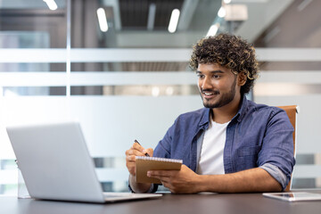 A young Indian man is sitting in an office at a desk, looking at a laptop screen and making notes...