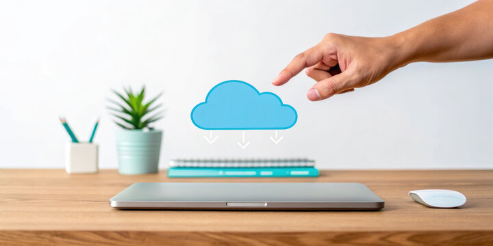 Cloud computing concept with hand pointing at blue cloud icon above closed laptop on wooden desk with plant and mouse - Powered by Adobe