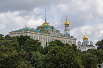 Fototapeta premium Grand Kremlin Palace and golden-domed cathedral complex in Moscow, framed by lush green trees under cloudy sky