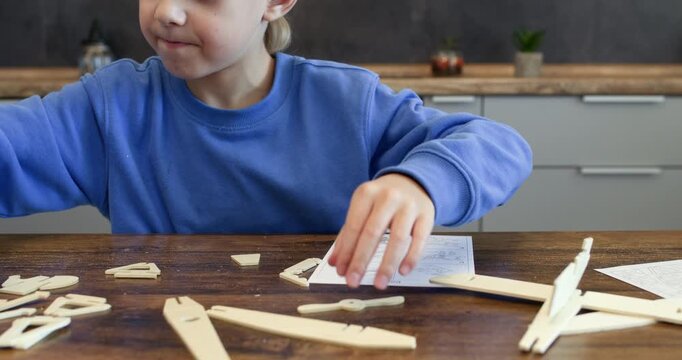 Engaged boy seeks wooden airplane detail looking at instruction guide at desk. Little schoolboy assembles vintage aeroplane of construction set in kitchen