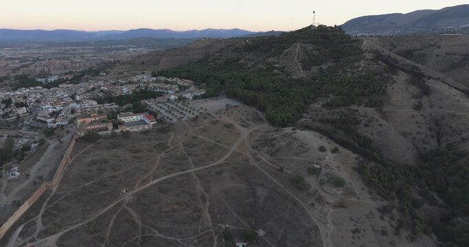 Drone aerial revealing Granada city from foothill ridge at sunrise, winding paths and warm dawn light near the Alhambra, Andalusia Spain
