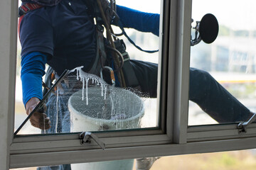 Professional window cleaner working on exterior glass of high-rise building using safety rope and bucket, maintenance service concept