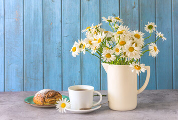 Summer breakfast still life; white cup of coffee, bun and bouquet of white daisy flowers on a table against blue paint wooden planks