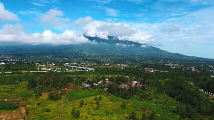 Mountain covered by clouds overlooking green farmland and rural settlement under blue sky, captured from a drone perspective.