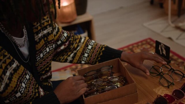 African American shop worker carefully arranging vintage eyeglasses and sunglasses on wooden display table with red sale sign on it in cozy secondhand shop with warm lighting and retro atmosphere