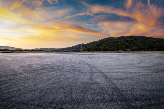 Dramatic empty asphalt ground with tire marks near a green mountain under a beautiful sunset sky.