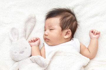 Top view of a baby sleeping on a bed. With a white doll rabbit. Beautiful portrait of a small asian baby. A cute little newborn baby in a blanket.