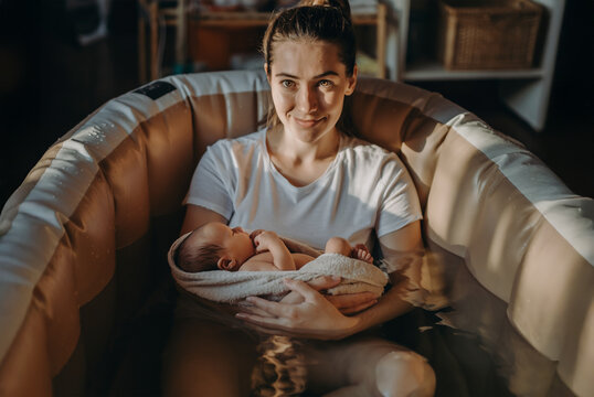 Woman sitting in birthing pool holding newborn baby wrapped in towel. Free birth. Home birth