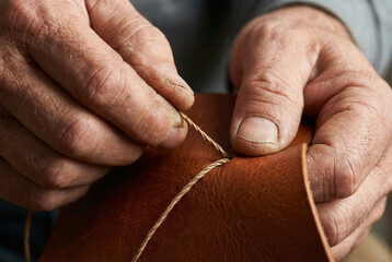 Extreme close-up of artisan hands sewing brown leather with waxed thread showing skin texture and detail