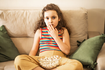 Little cute girl kid watching TV movie on sofa in the living room with popcorn