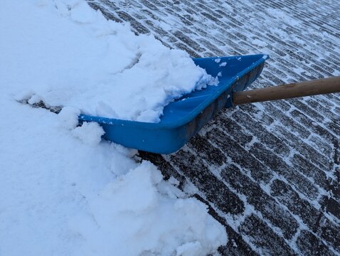 A bright blue plastic shovel sits on a snow-covered pavement, ready for shoveling after a snowfall. The contrast highlights winter cleanup, cold air, and the effort of clearing driveways and paths.