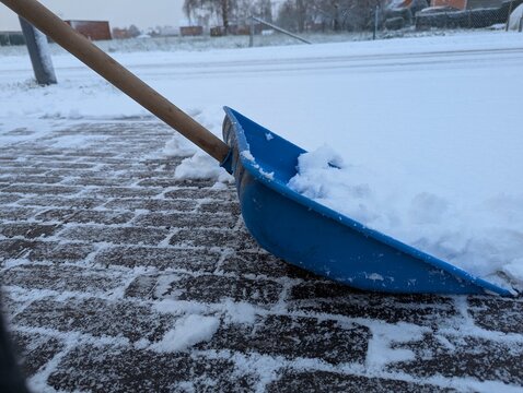 A blue plastic bright shovel sits on a snow-covered pavement, ready for shoveling after a snowfall. The contrast highlights winter cleanup, cold air, and the effort of clearing driveways and paths.