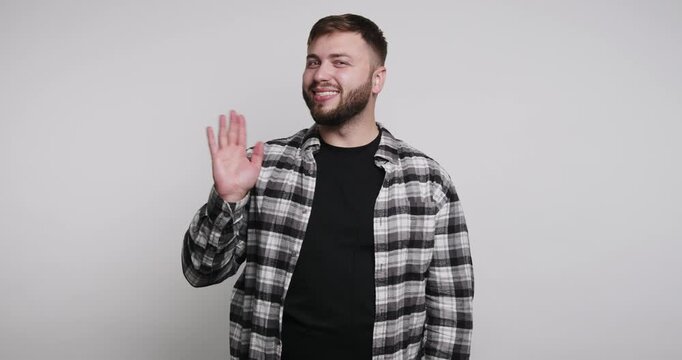 Man waves hello in a simple setting with a plain background and casual clothing while smiling and looking happy