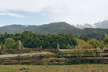 Fototapeta premium Fence, meadow, forest, mountains, landscape, rural wooden fence with grassy meadow and forest leading to distant mountain peaks under blue sky