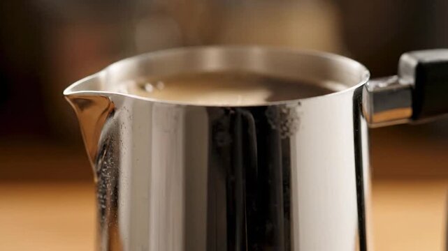 A stainless steel pitcher with coffee being poured inside; a close-up shot with brown background