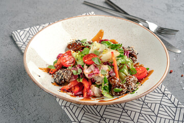 Asian-inspired salad with glazed teriyaki beef chunks in sesame seeds, fresh mixed greens, cherry tomatoes, carrot strips and soy dressing in rustic white bowl. Vibrant fusion gourmet dish
