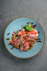 Sliced rare roast beef salad with roasted baby potatoes, fresh greens, red onion rings, cherry tomatoes and balsamic glaze in light blue bowl. Gourmet beef salad, elegant restaurant-style, top view