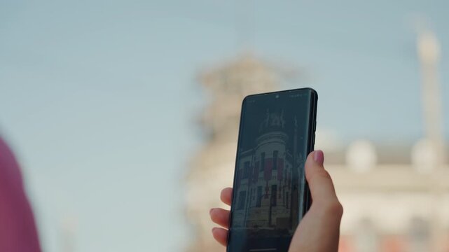 hand holding smartphone capturing historic facade against clear blue sky with decorative string lights, ornate building under scaffolding, closeup of persons hand with painted nails, soft bokeh