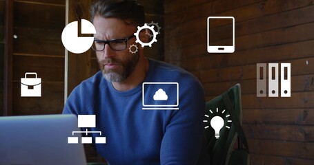 Working bearded man wearing glasses, blue sweater at laptop in wood room with green jacket, icons