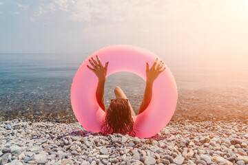 Beach woman inflatable sitting on a pebble shore while holding a pink ring over her head during a golden sunset