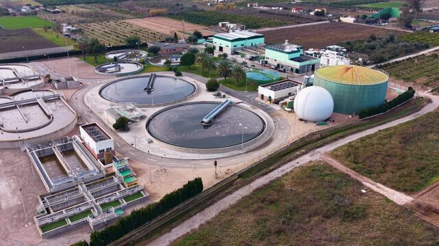 Orbiting aerial of a Spanish wastewater treatment plant with circular clarifiers, sludge processing units and biogas storage, showcasing modern sewage and water recycling infrastructure