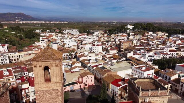 Aerial flight above the historic Santa Maria monastery, revealing the dense rooftops of El Puig and the surrounding Valencian countryside under a bright Mediterranean sky