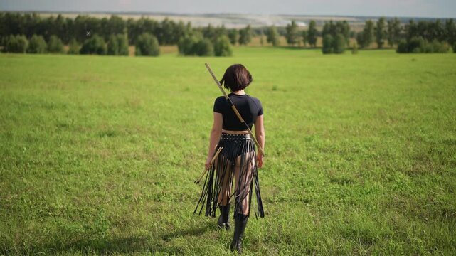 woman walking across green field, solitary leatherclad warrior carrying spear on back, slow steady stride toward distant treeline under soft sunlight, cinematic atmosphere of resilience and quiet