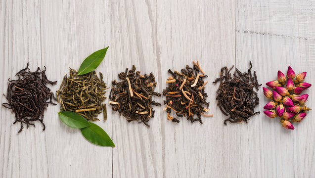 Assorted loose tea types and dried rosebuds arranged in a row on wooden background. Variety of herbal and black tea blends.