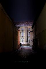 Ciemna, mroczna uliczka, zaułek. W zaułku panują ciemności. Na wprost widać dobrze oświetlony, zabytkowy budynek.