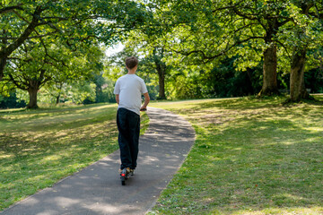 Fototapeta premium Boy rides electric scooter along path in green park during sunny day